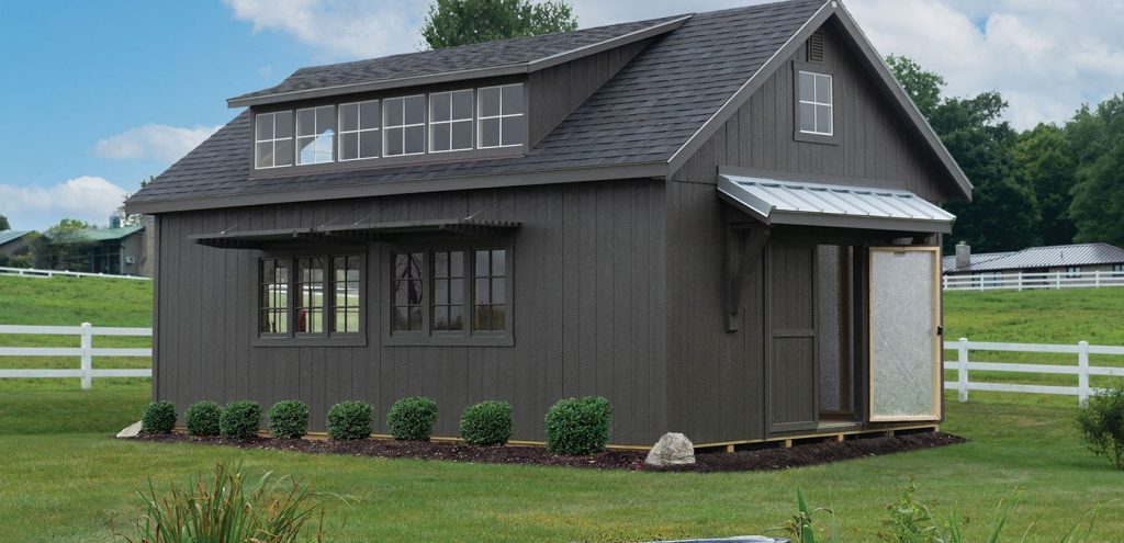 Dark gray storage shed with dormer windows and metal awning on a landscaped lawn